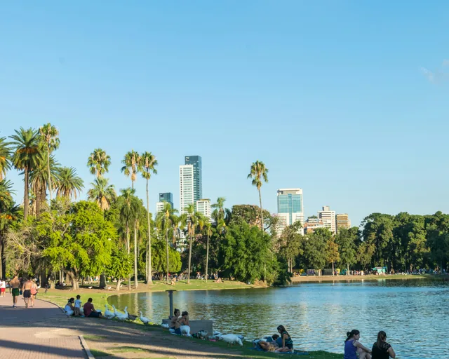 People sitting beside a lake in a Buenos Aires park, with palm trees and towers beyond.