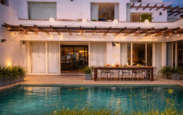 The villa pool at dusk, with a long outdoor dining table and string lights under the pergola.