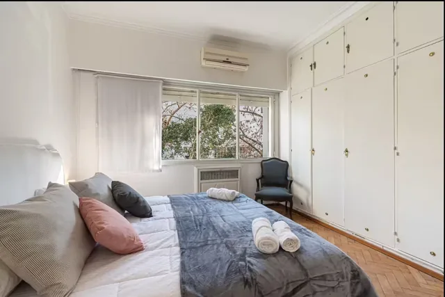 A private double bedroom with white wardrobes, a large window and wood flooring.