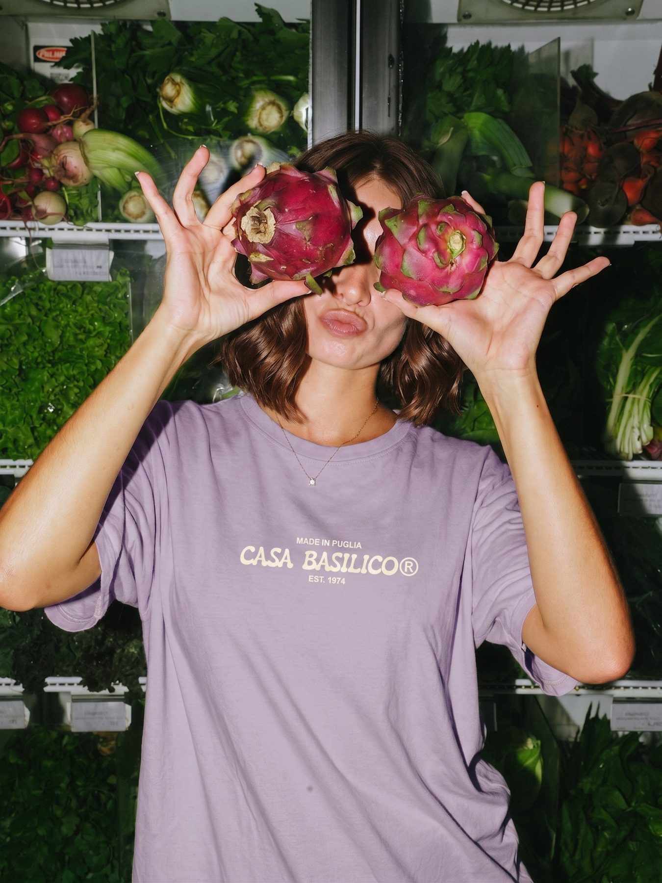 Woman in the lilac Casa Basilico tee holding two dragon fruits over her eyes in front of a vegetable fridge