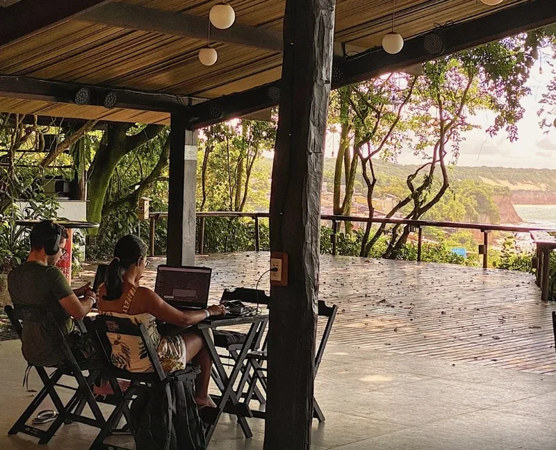 Two guests working on laptops at a table on the covered open-air deck, with a sea and clifftop view through the trees
